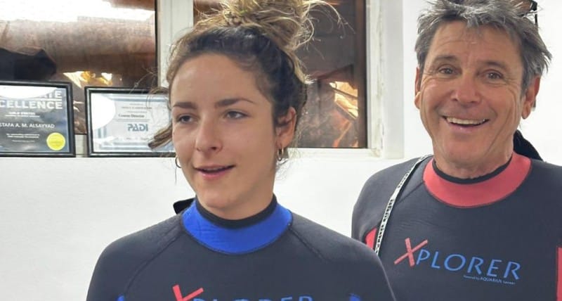 Two divers in Xplorer wetsuits pose indoors at a diving center with certificates on the wall, smiling at the camera.