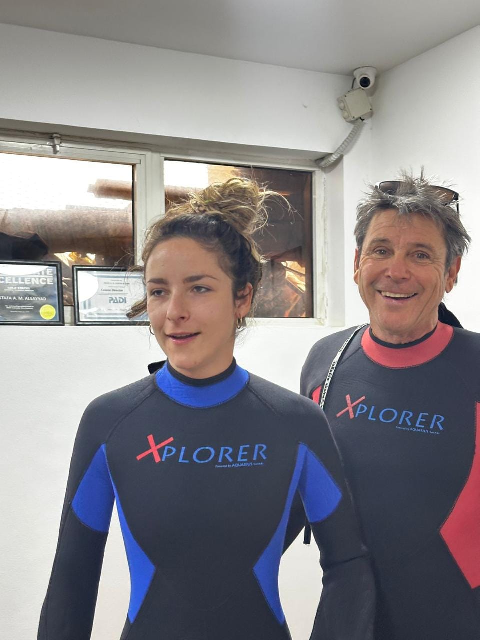 Two divers in Xplorer wetsuits pose indoors at a diving center with certificates on the wall, smiling at the camera.